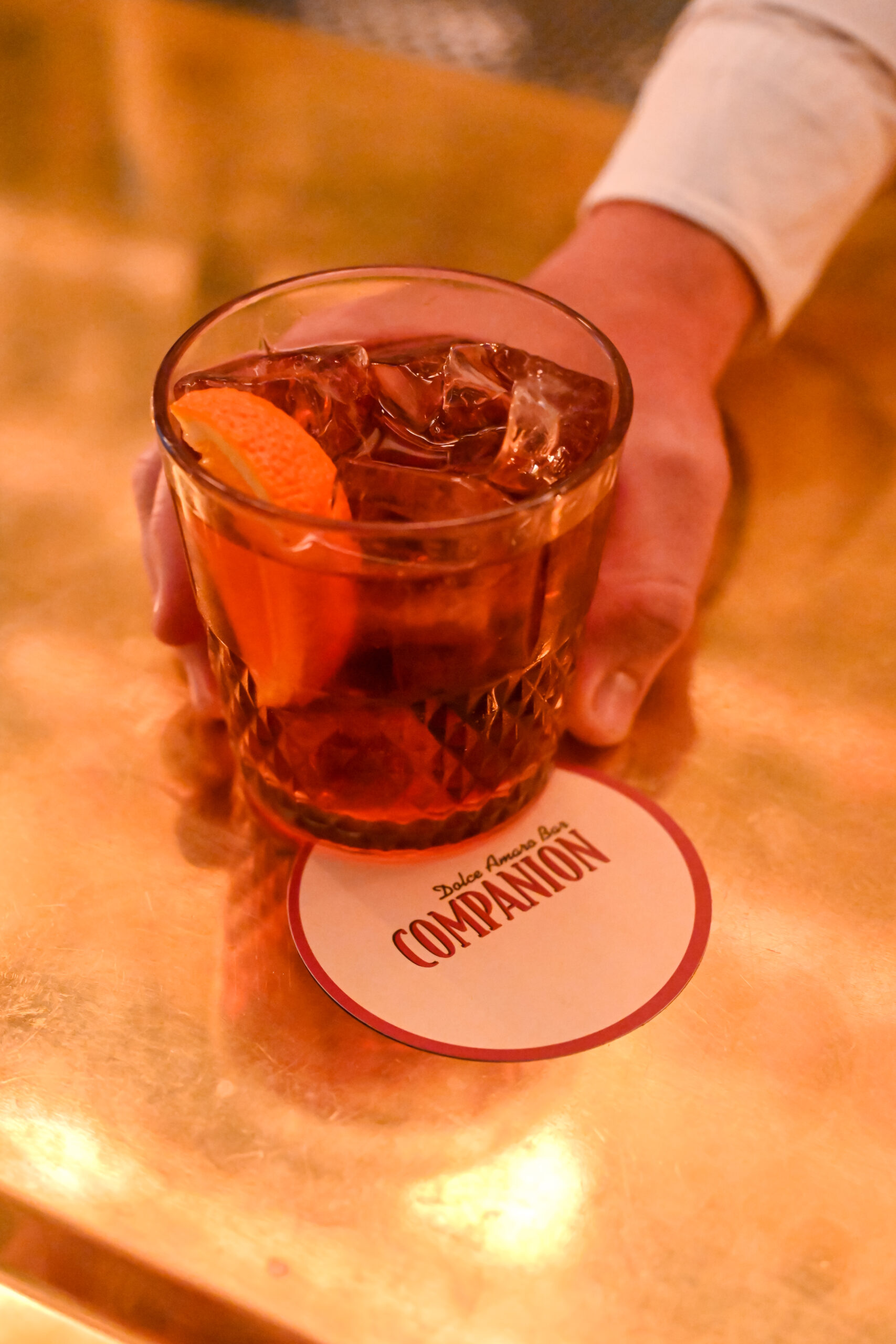 A drink with icecubes and an orange peel sitting on a coaster with the logo Companion Dolce Amaro Bar on it.